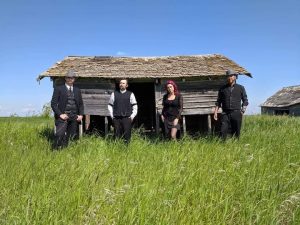 Four band members stand in long grass in front of a decaying building